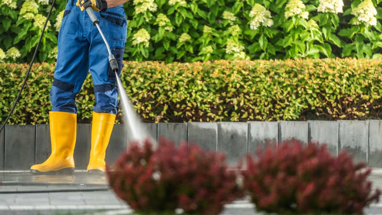 A person in yellow boots pressure washing a driveway behind two red bushes